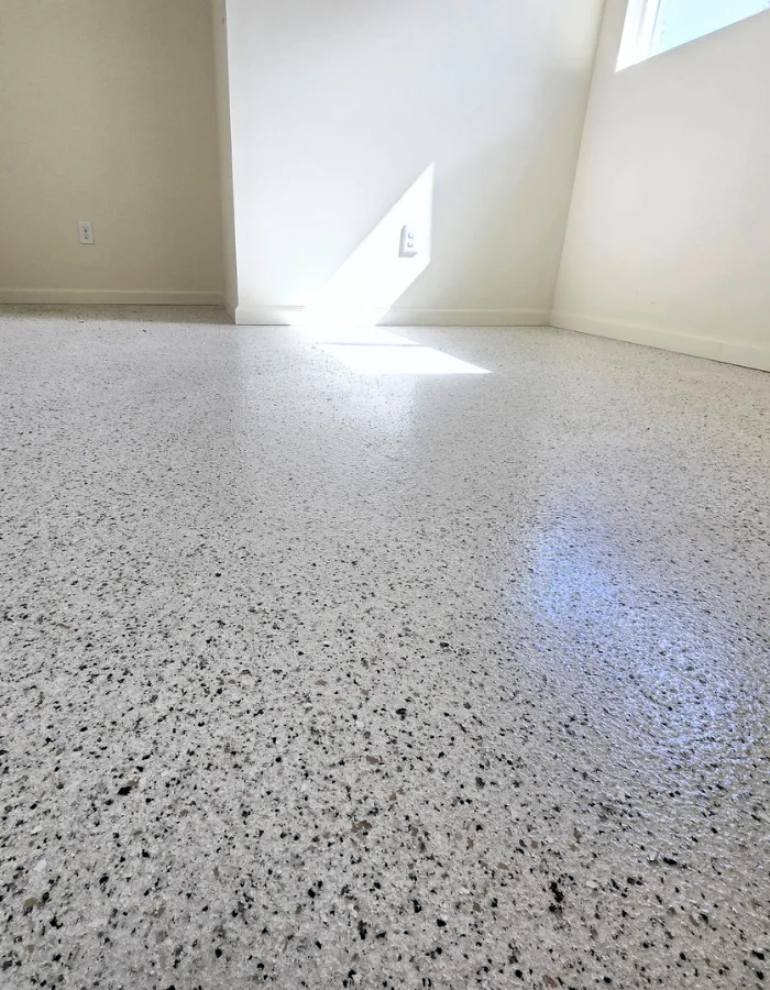Empty sunlit corner of a white room with a speckled terrazzo floor; a triangular patch of sunlight shines on the wall near an outlet.