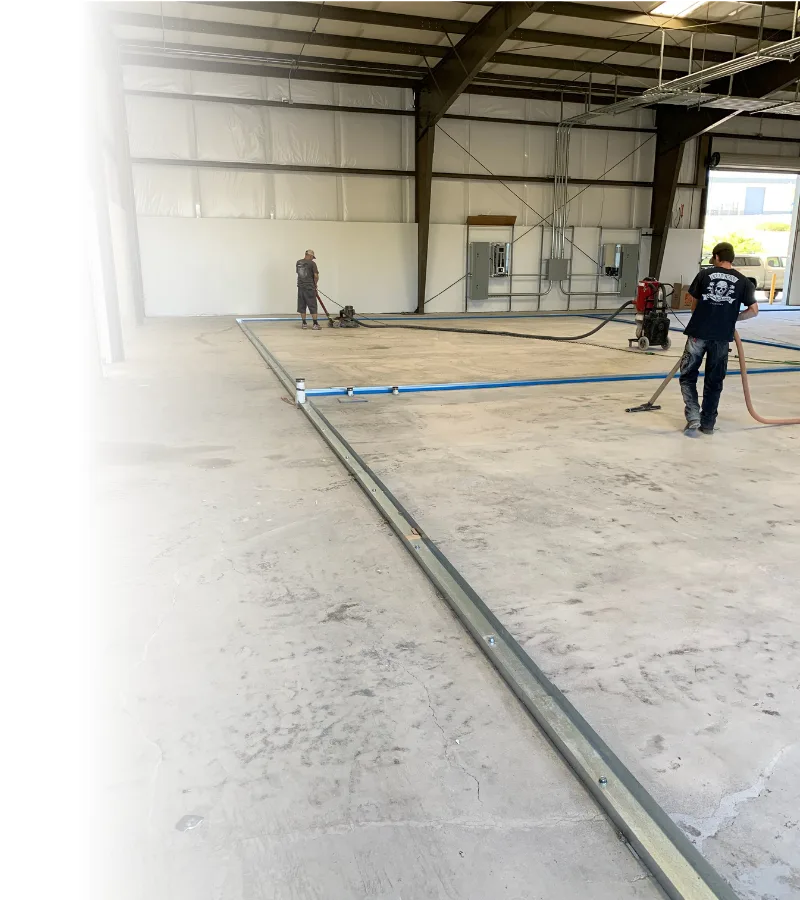 Two workers in a large industrial warehouse lay down metal rails and hoses on a dusty concrete floor.