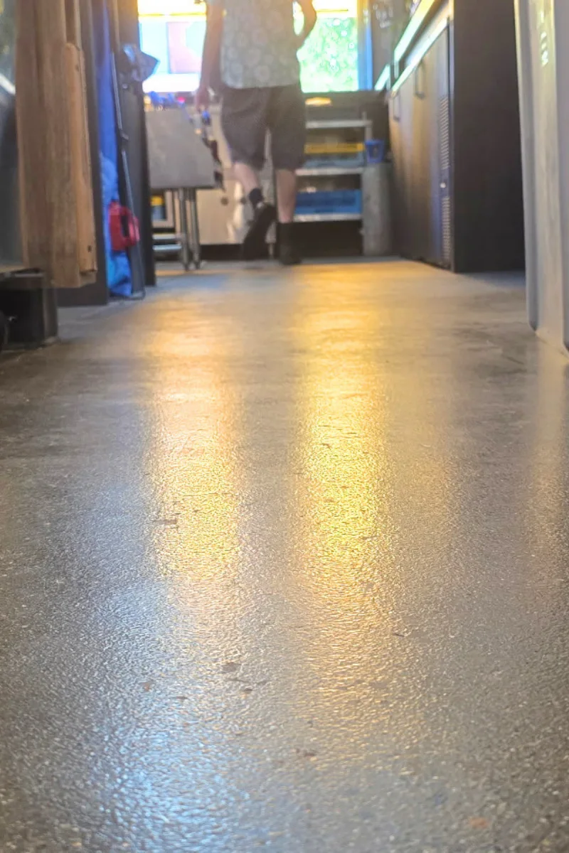 Person walking away down a narrow indoor hallway with equipment on the left and a cart visible at the far end, bathed in warm overhead light.